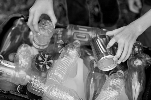 A close-up black and white image showing a person's hands sorting through a collection of recyclable waste, primarily consisting of empty plastic bottles and a metal tin can. The plastic bottles vary in shape and size, with some capped and others without lids, and display textured surfaces typical of PET material. The metal tin can is held by one hand, with a smooth, reflective surface and a slightly recessed top edge. The waste items are arranged on a flat surface, with some bottles leaning or stacked against each other, and the background is blurred. The image suggests activity related to waste sorting or recycling, which is consistent with rubbish removal services offered by Commercial Waste Removal Cranford, located near Cranford and the TW postcode area. The scene emphasizes environmental responsibility through recycling, with a focus on waste segregation and disposal in a professional context. The lighting is even, highlighting the textures and details of the bottles and can, reinforcing the theme of waste management and recycling logistics.