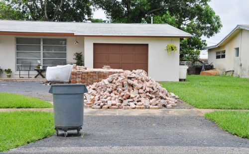 A pile of broken bricks and rubble, situated on a paved driveway in front of a detached single-storey building with a brown garage door, white walls, and a large window with blinds. The rubble consists of various shades of light brown and beige bricks, with some bricks still partially stacked beside the pile. To the left of the rubble, a grey wheelie bin is positioned on the driveway, and a white cylindrical object, possibly a rolled-up construction material, is placed on top of a small brick wall. The driveway is bordered by neat, green grass on both sides, leading up to the building which has a small garden area with a few plants visible near the window. In the background, there are trees and another property with a similar single-story structure, indicating a residential area. This scene depicts a typical scene for rubbish removal services, such as those offered by Commercial Waste Removal Cranford, in a suburban setting near Cranford.