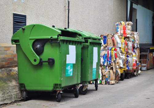 A green wheeled rubbish bin with a hinged lid, positioned on a paved surface against a beige building wall, with two white labels on the front. To the right of the bin, there are multiple tightly stacked bundles of flattened cardboard boxes secured with strips of tape, varying in size and color, including brown, blue, red, and white. The cardboard bundles are situated on the ground next to the bin, near a large window with reflective glass and a dark frame, suggesting an urban or industrial environment, possibly in Cranford or nearby. This scene exemplifies typical waste collection and disposal setup for commercial or residential properties, as part of waste management services such as those provided by Commercial Waste Removal Cranford.