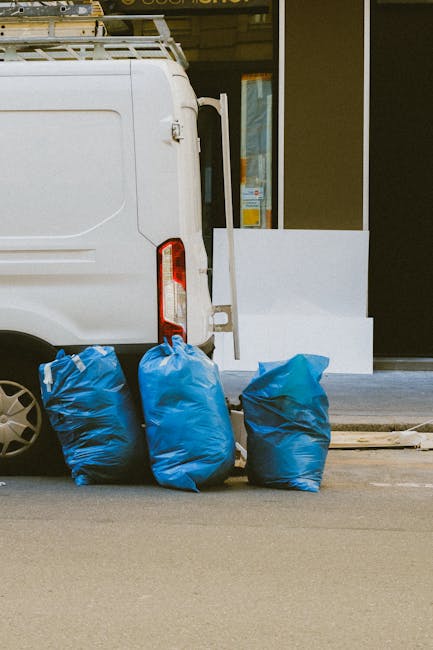 Piles of black rubbish bags and discarded materials, including a piece of cardboard and some wooden planks, are stacked in front of a red metal door on an exterior wall. The door is decorated with gra
