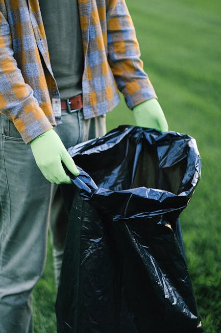 A person wearing a yellow and blue checkered shirt, a green cardigan, and light grey trousers is holding a large black plastic rubbish bag open with both hands. The individual is wearing bright green